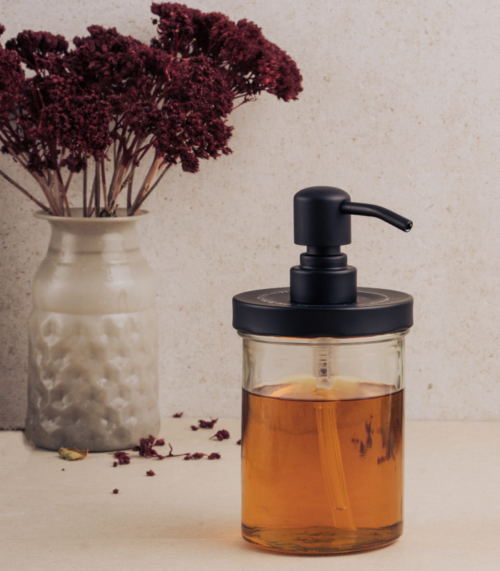 Mangolds Pumpspender-Deckel auf Glas mit brauner Flüssigkeit vor Vase mit roten Strauchblüten